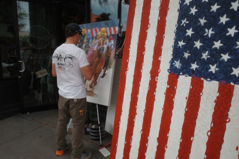 A street artist painting a horse-racing scene at an easel on the sidewalk outside a shop, beside a large textured painting of an American flag.