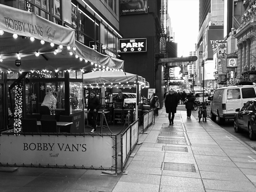 Black-and-white street scene of string-lit awnings and outdoor seating at Bobby Van's Grill along a busy city sidewalk.