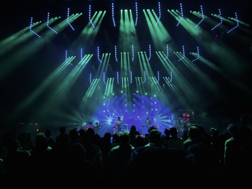 Goose playing on stage at SPAC, with green and blue light beams fanning out over a silhouetted crowd.