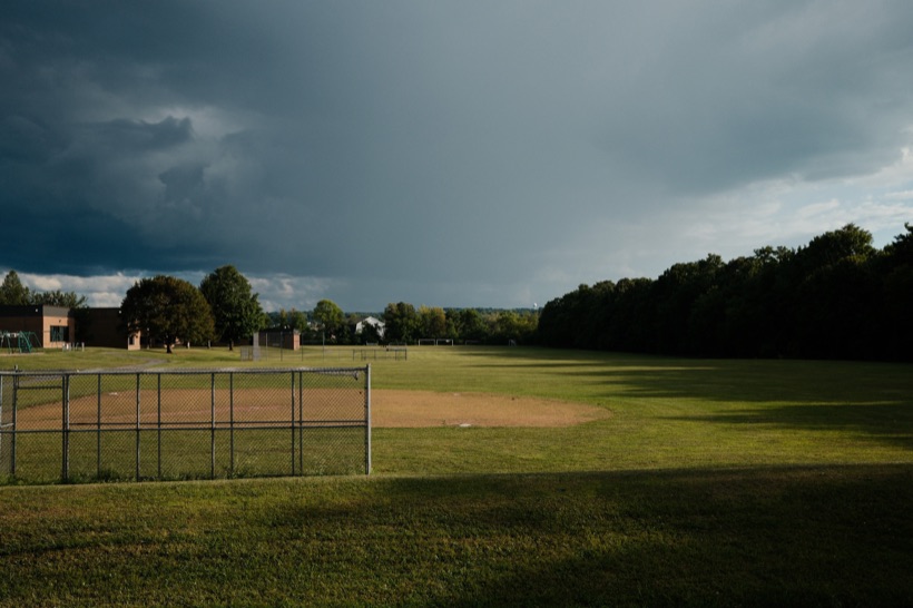 Dark storm clouds building over a school's ball fields, with late sun lighting the grass, a chain-link backstop, and the infield dirt.