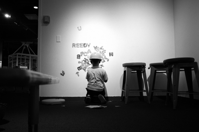 Black-and-white photo of a small child in a white hard hat kneeling at a wall of magnetic letters at the MOST, Syracuse's Museum of Science & Technology.