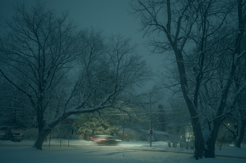 A car's taillights blur past a stop sign on a snowy street at night, under bare trees heavy with snow.