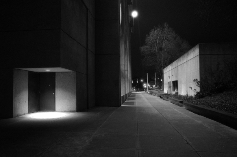 Black-and-white night view down a long concrete walkway between campus buildings at Syracuse University, a doorway lit by a single light.